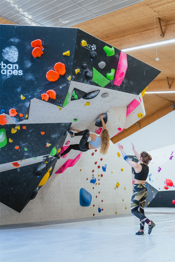 Eine Frau hängt an der Boulderwand. Eine Frau steht mit gestreckten Armen hinter ihr, um sie zu sichern.