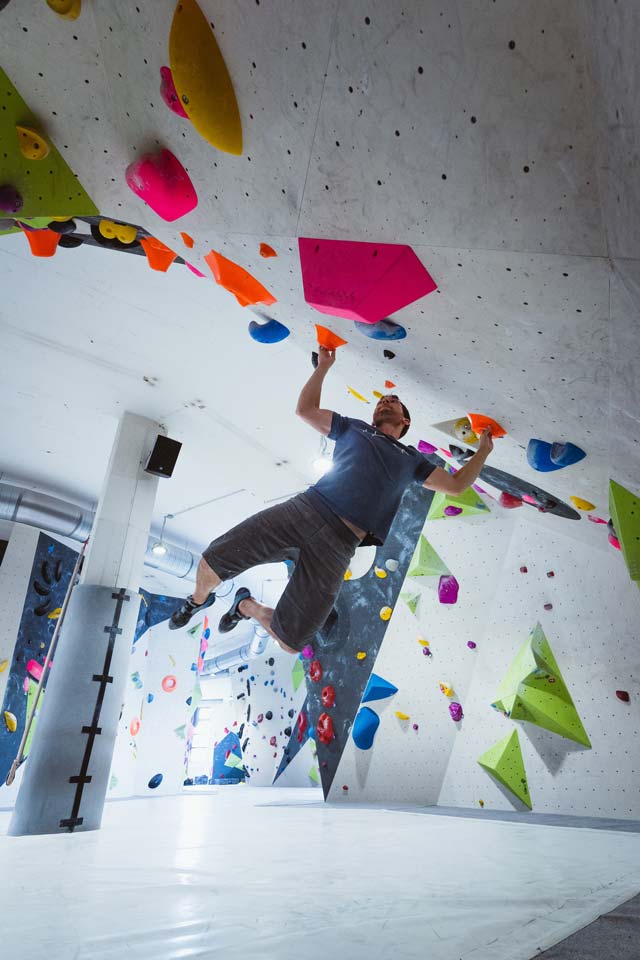 Boulderer in blauem Shirt beim Sprung, hält sich an zwei orangen Griffen fest