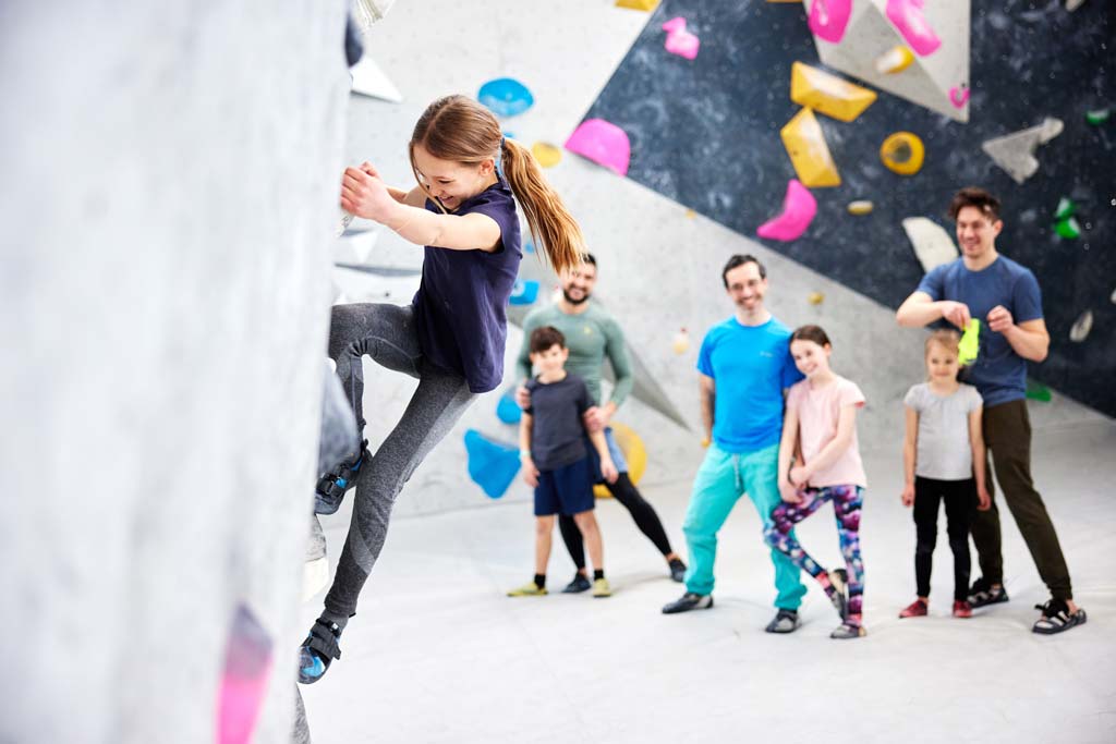 Mädchen an der Boulderwand, im Hintergrund andere Kinder mit ihren Vätern.