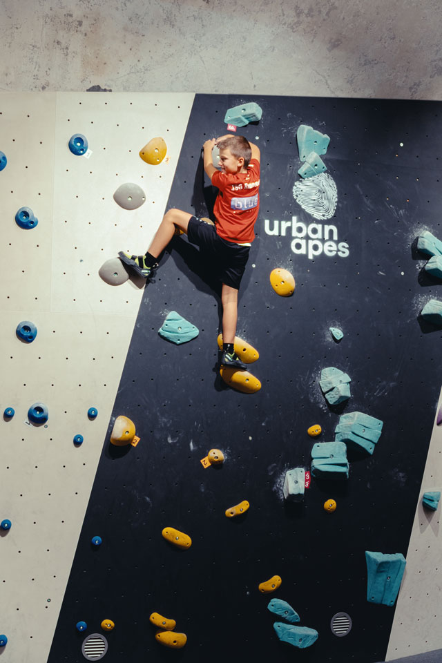 Junge in rotem Shirt klettert an hoher Boulderwand mit gelben und blauen Griffen.